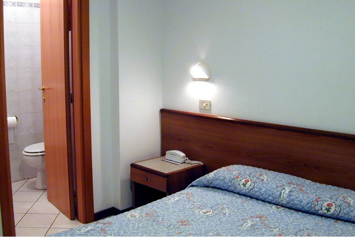A bedroom with a wooden headboard, a bedside table with a phone, a wall-mounted light, and a floral-patterned bedspread.