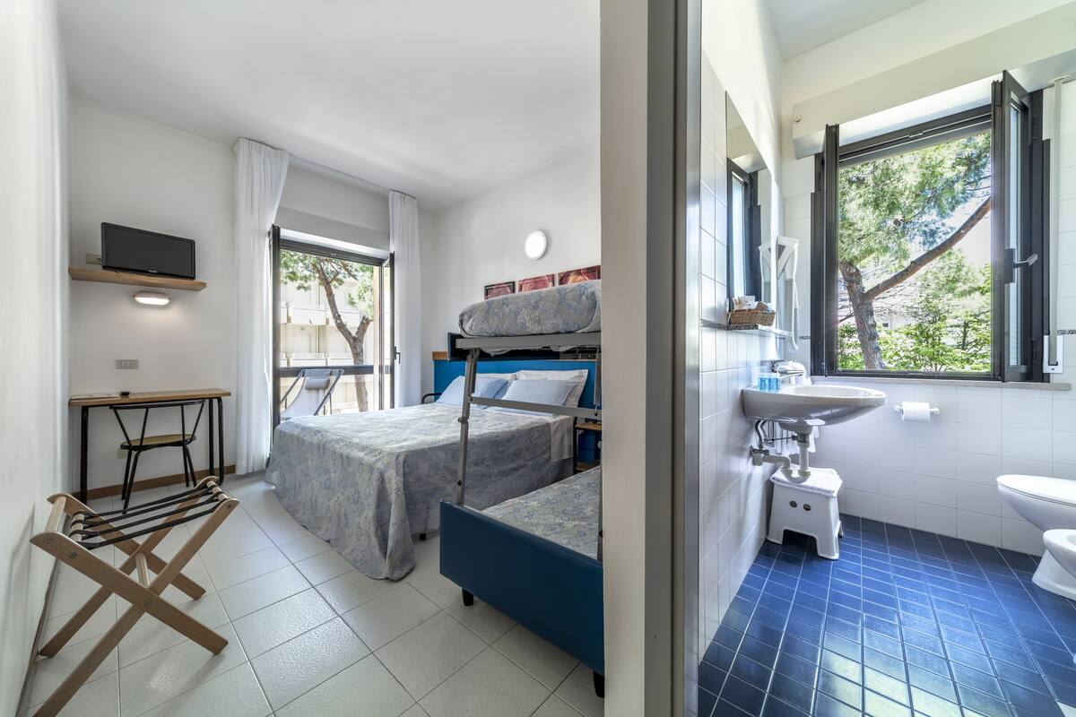 A room with two beds, a desk, a TV, a window with a view of trees, and a bathroom with a sink and toilet.