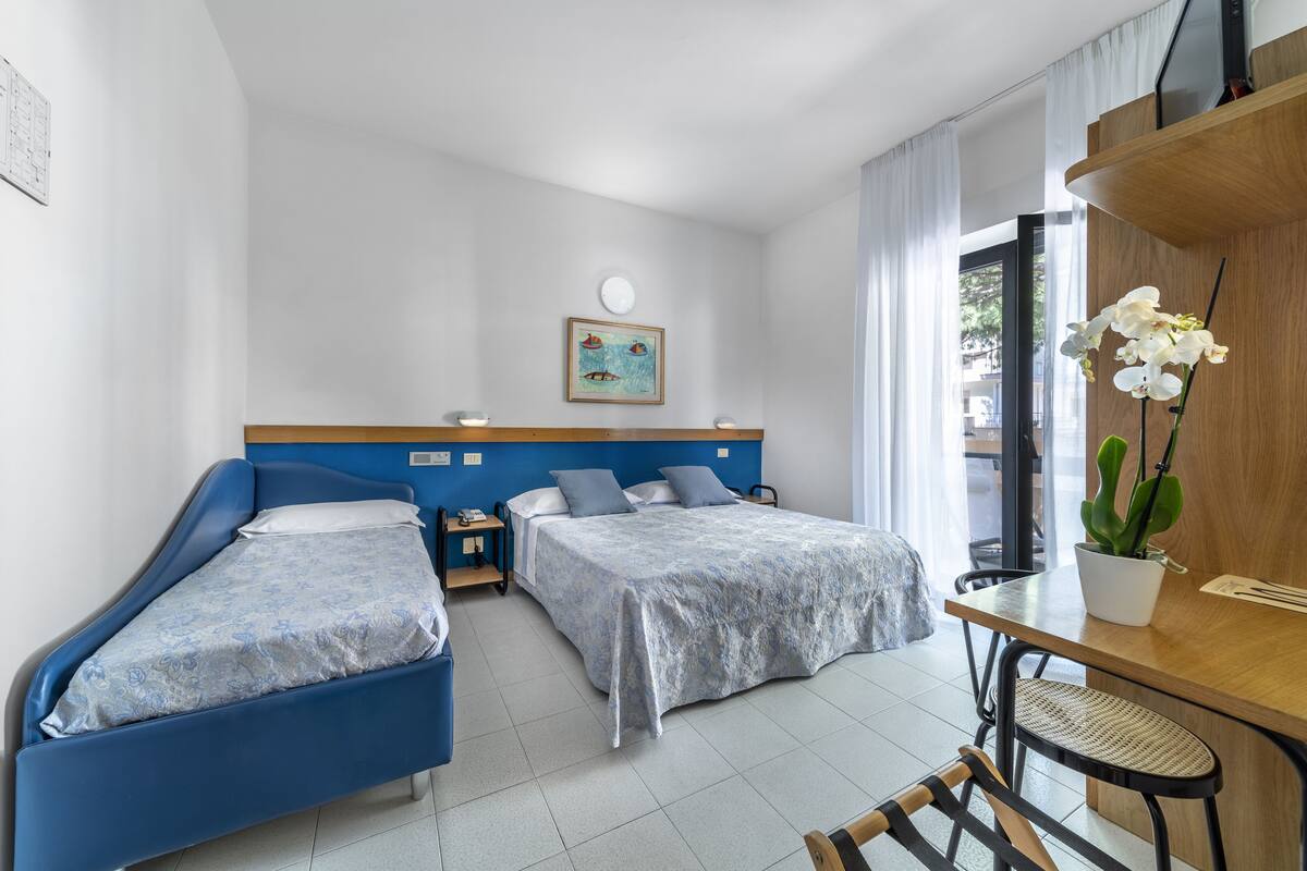 A hotel room with two beds, a desk, a chair, a potted plant, and a window with curtains.