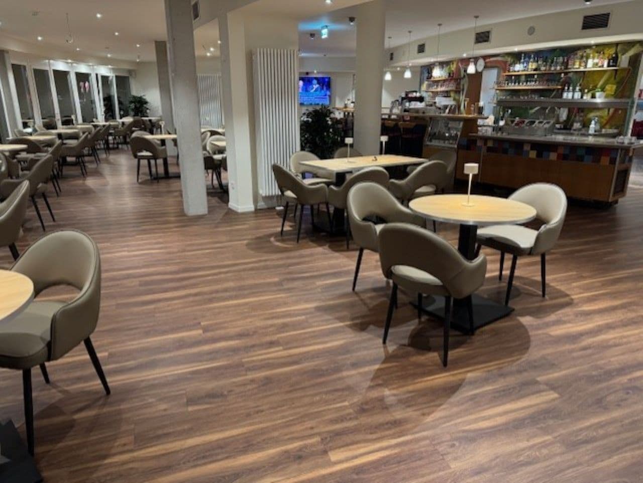 A modern restaurant with wooden flooring, round tables, and cushioned chairs.