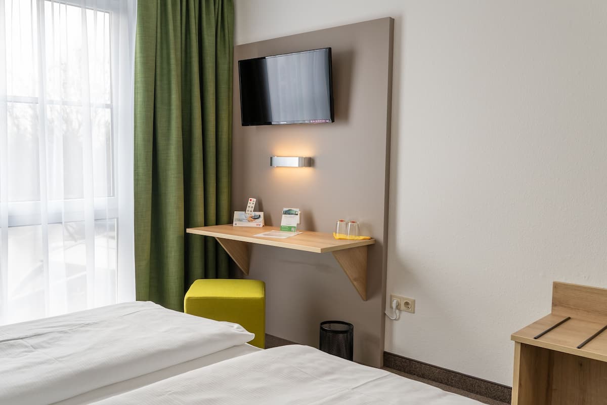 A hotel room with a bed, a desk with a flat-screen TV, a small trash bin, and a wooden nightstand.