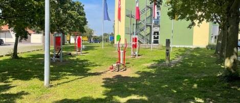 Un área verde con juegos infantiles, un asta de bandera y un edificio de fachada verde.