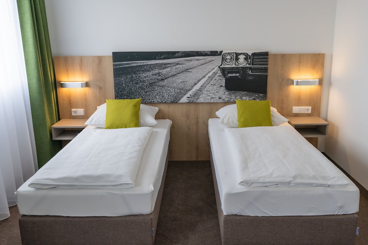 A twin room with two beds, a wooden headboard, bedside lamps, and a road painting on the wall.