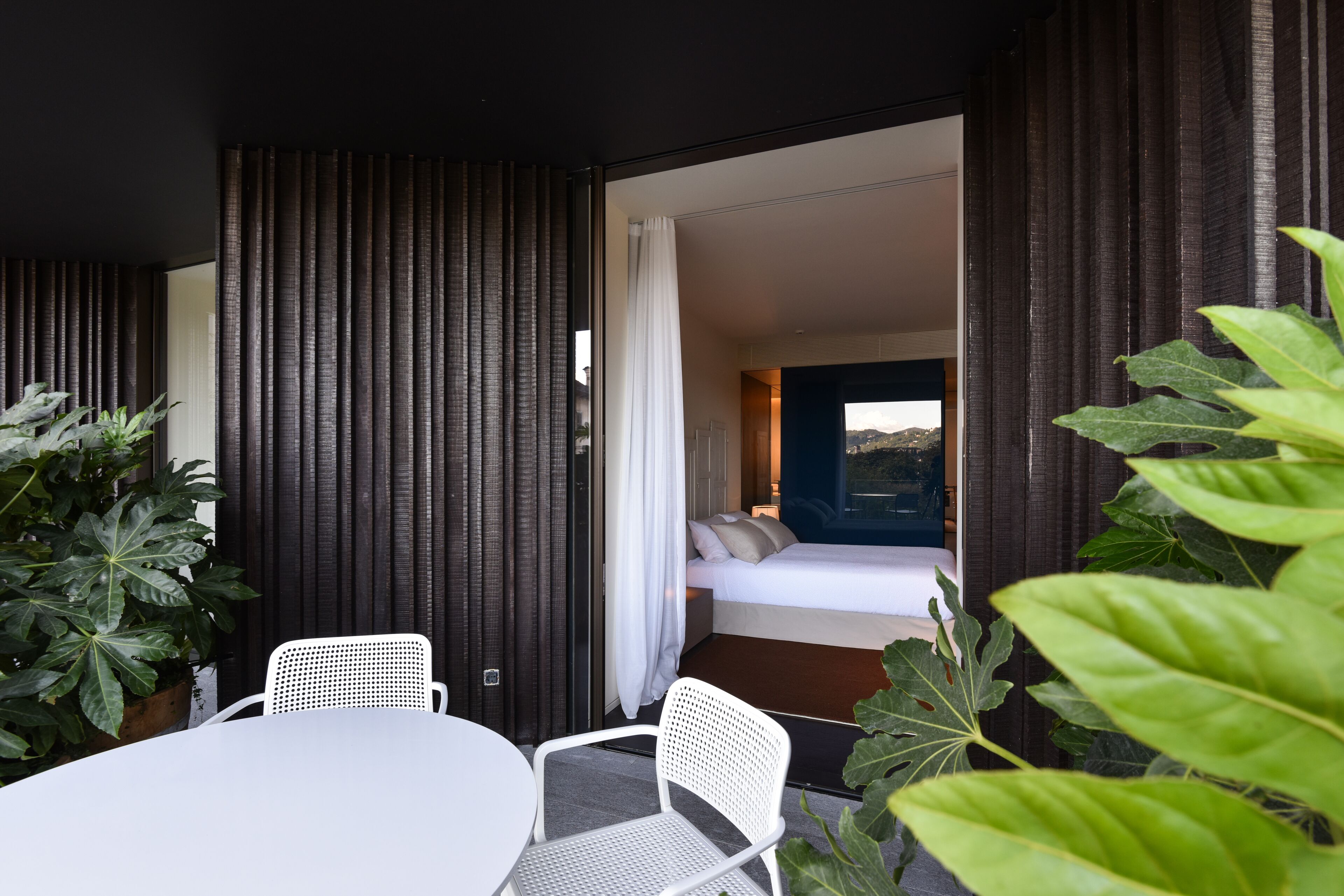 A modern hotel room with a bed, a small table, and chairs. There is a large window with curtains, and the room has a dark wall and a plant.