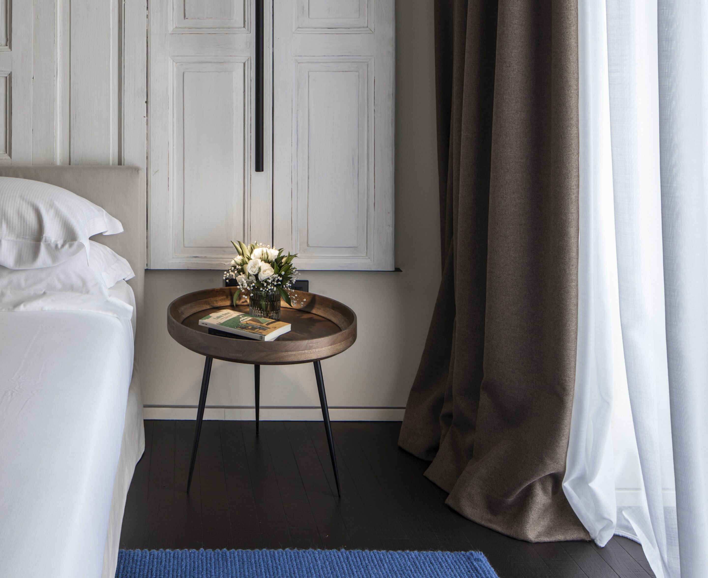 Un lit bien fait, avec du linge de lit blanc, une petite table ronde en bois sur laquelle se trouvent un livre et un vase de fleurs, et un plancher en bois foncé.