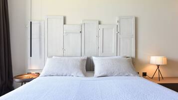 En pænt redt seng med hvide sengetæpper, et natbord med en lampe, og en garderobe med hvide døre.
