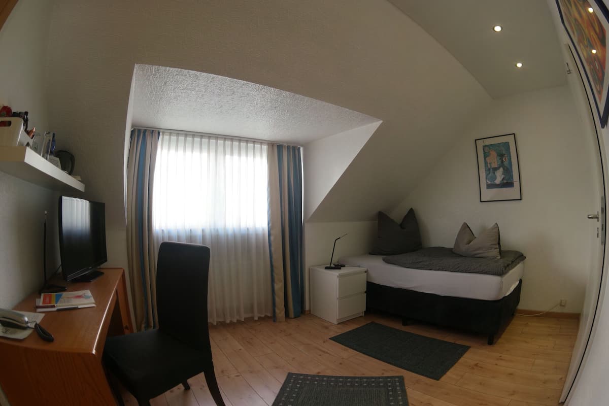 Un dormitorio con cama, escritorio con computadora, silla, ventana con cortinas y un cuadro en la pared.