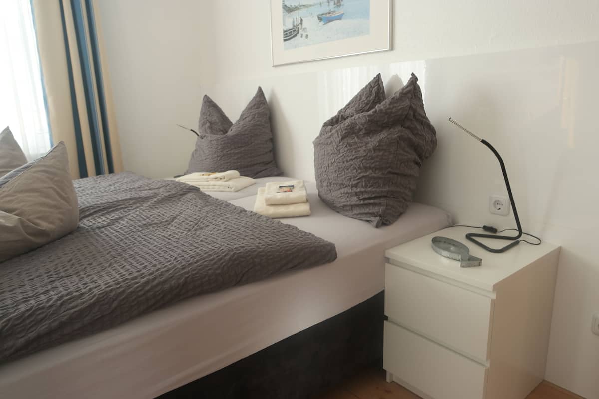 Una cama bien hecha con ropa de cama y almohadas grises, una mesita de noche con una lámpara y un teléfono, y un cuadro en la pared.