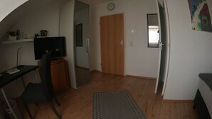 A room with a wooden floor, a desk with a chair, a television, a mirror, a door, and a rug. - Hotel Merlin (Cologne)