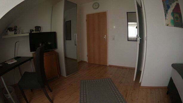 A room with a wooden floor, a desk with a chair, a television, a mirror, a door, and a rug. - Hotel Merlin (Cologne)