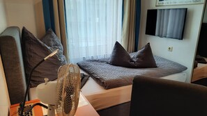 A hotel room with a bed, a desk, a chair, a standing fan, and a TV mounted on the wall. - Hotel Merlin (Cologne)