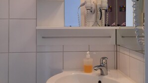 A bathroom with a white sink, a mirror, a hairdryer, and a shelf with toiletries. - Hotel Merlin (Cologne)