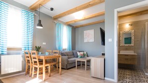 A modern living room with a wooden dining table, grey sofa, and a small coffee table. There is a large window with blue curtains, a ceiling fan, and a mirror on the wall. - Domki  Polanki 23 (Wicko)