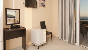 Habitación compacta con cama, escritorio con computadora, refrigerador pequeño, una silla y puerta corrediza de vidrio que da a un balcón con vista al mar.