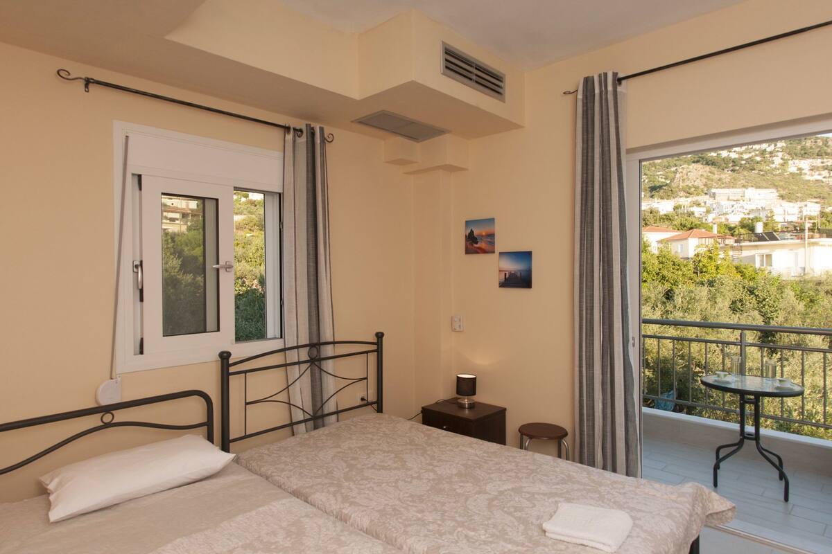 A bedroom with a bed, a bedside table, a small table with a lamp, and a view of a balcony with a table and chairs.
