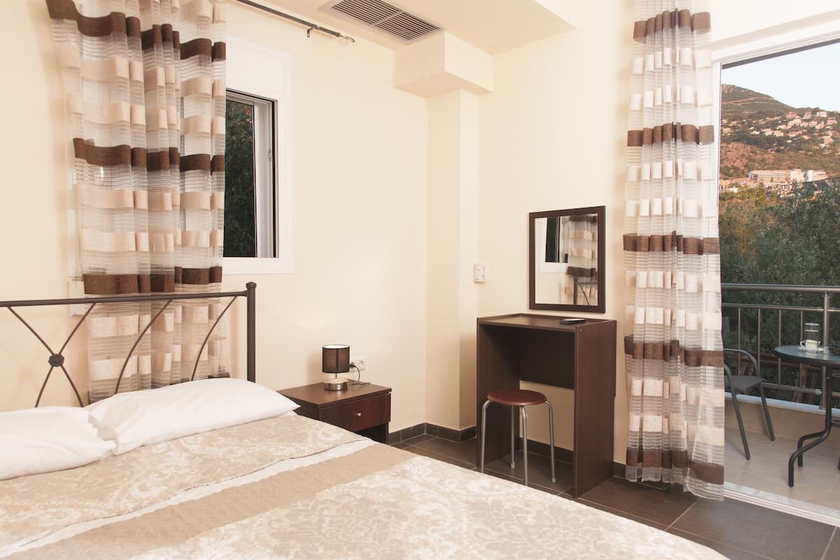 A bedroom with a bed, a desk, a chair, a window, and a balcony with a view of the hills.