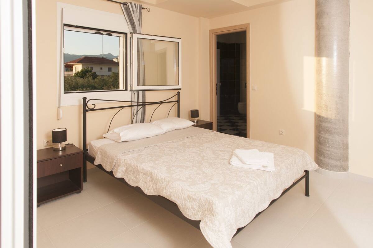 A neatly made bed with a white duvet, a bedside table with a lamp, and a window with a view of buildings and mountains.
