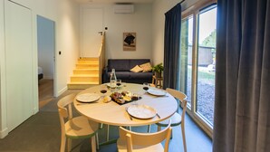 Een eetgedeelte met een ronde tafel, stoelen en een kroonluchter. Een trap leidt naar een andere kamer, een bank met kussens en een raam met gordijnen.