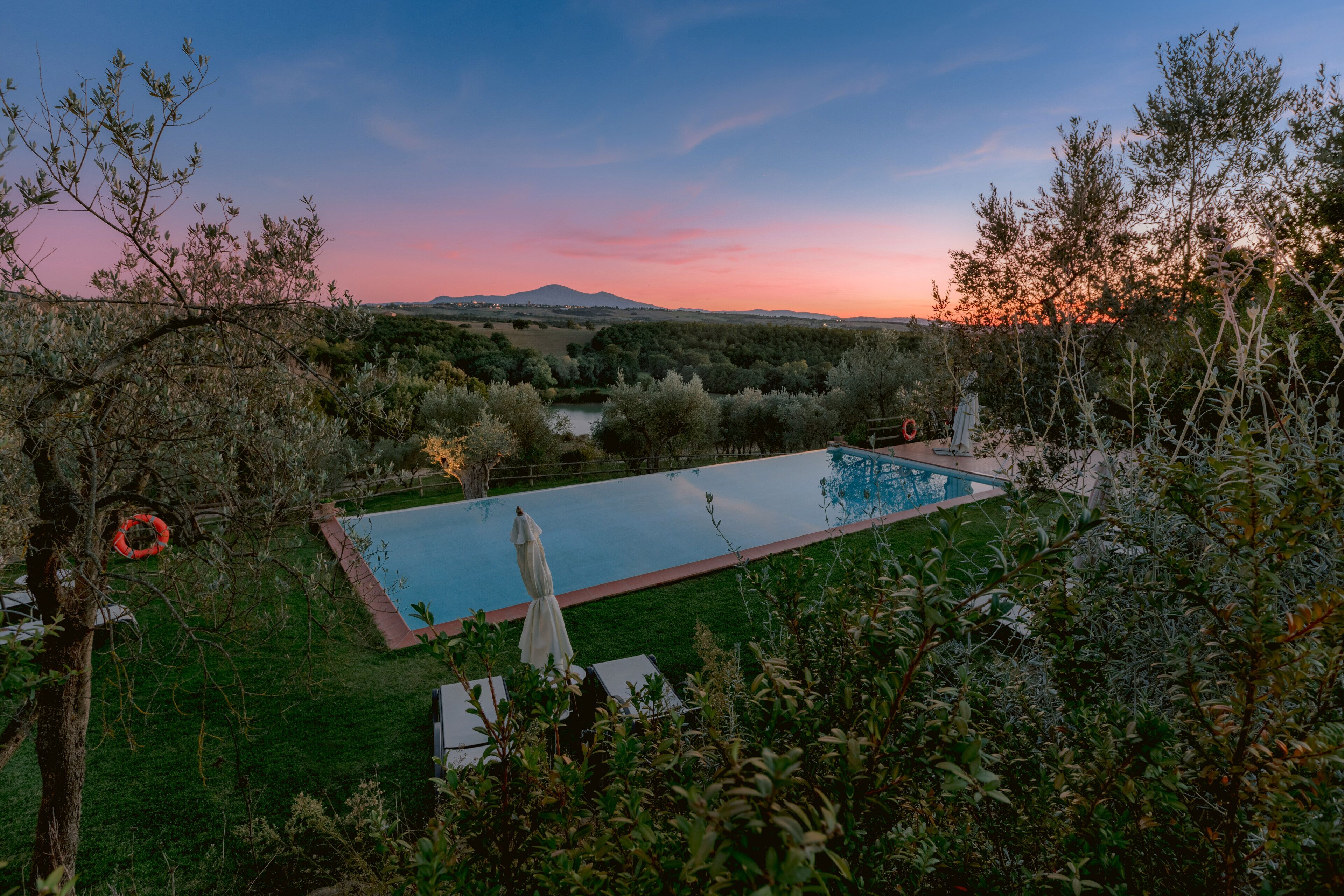 A rectangular swimming pool with lounge chairs and umbrellas, surrounded by greenery and trees, with a view of distant hills and a sunset sky.