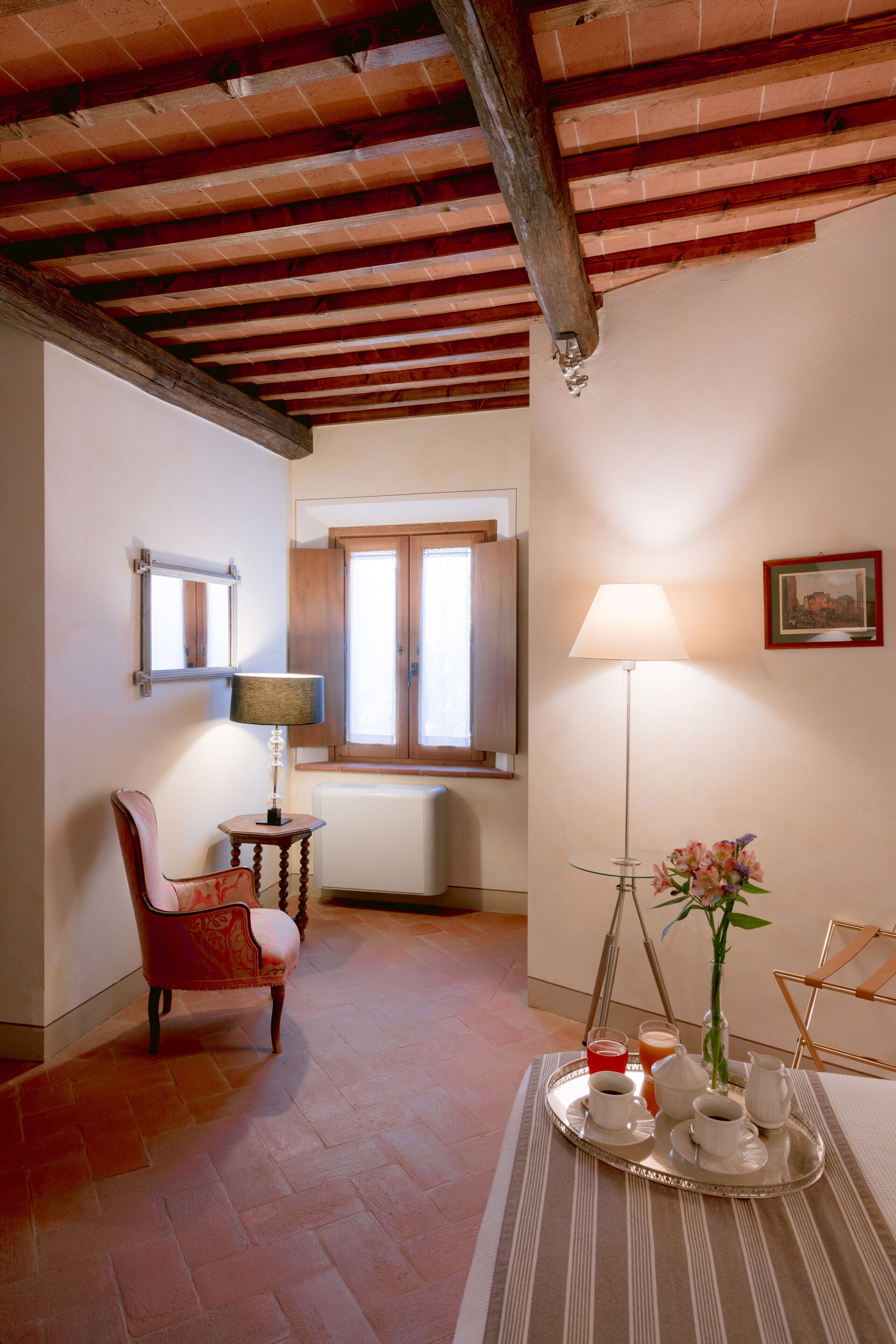 Una habitación acogedora con techo de madera, una silla, una mesa con una bandeja de desayuno, una lámpara y una ventana con contraventanas de madera.