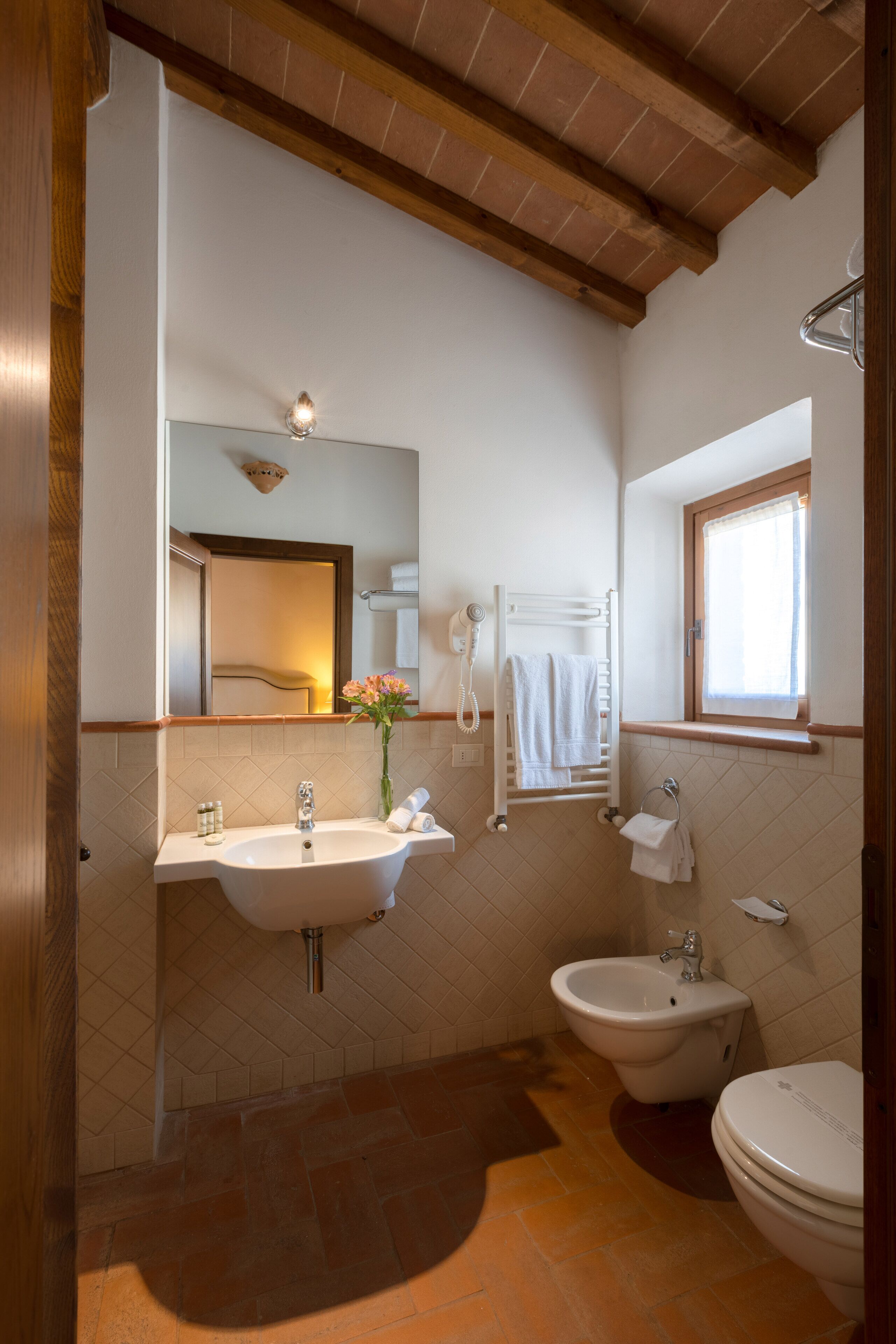 Ein Badezimmer mit Waschbecken, Bidet und Toilette, Holzbalken an der Decke und einem Fenster mit Handtuchhalter.