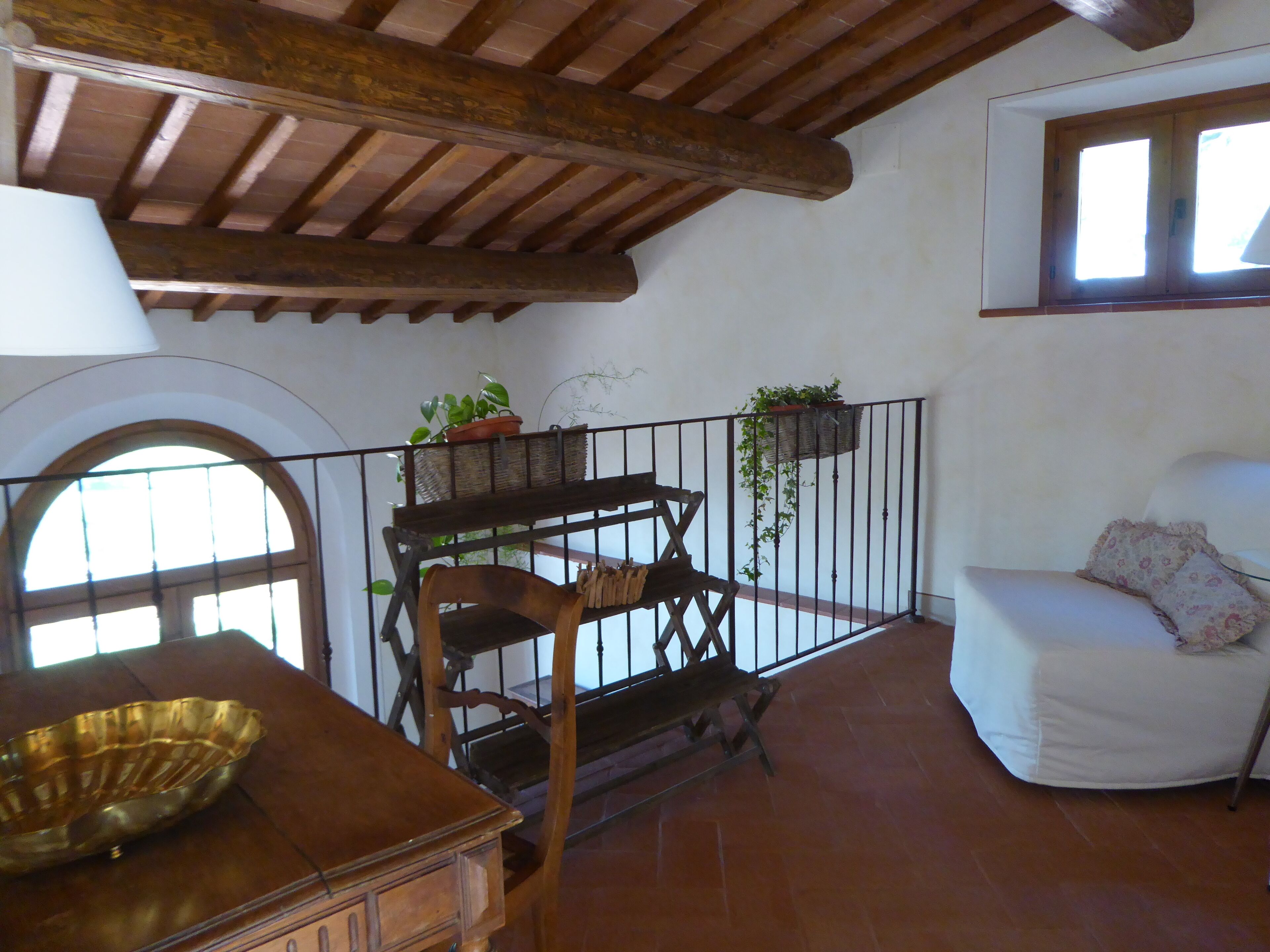 Una habitación rústica con un banco de madera, un sofá blanco y una mesa con un cuenco de latón. Hay plantas en macetas sobre una barandilla y una ventana con vistas.