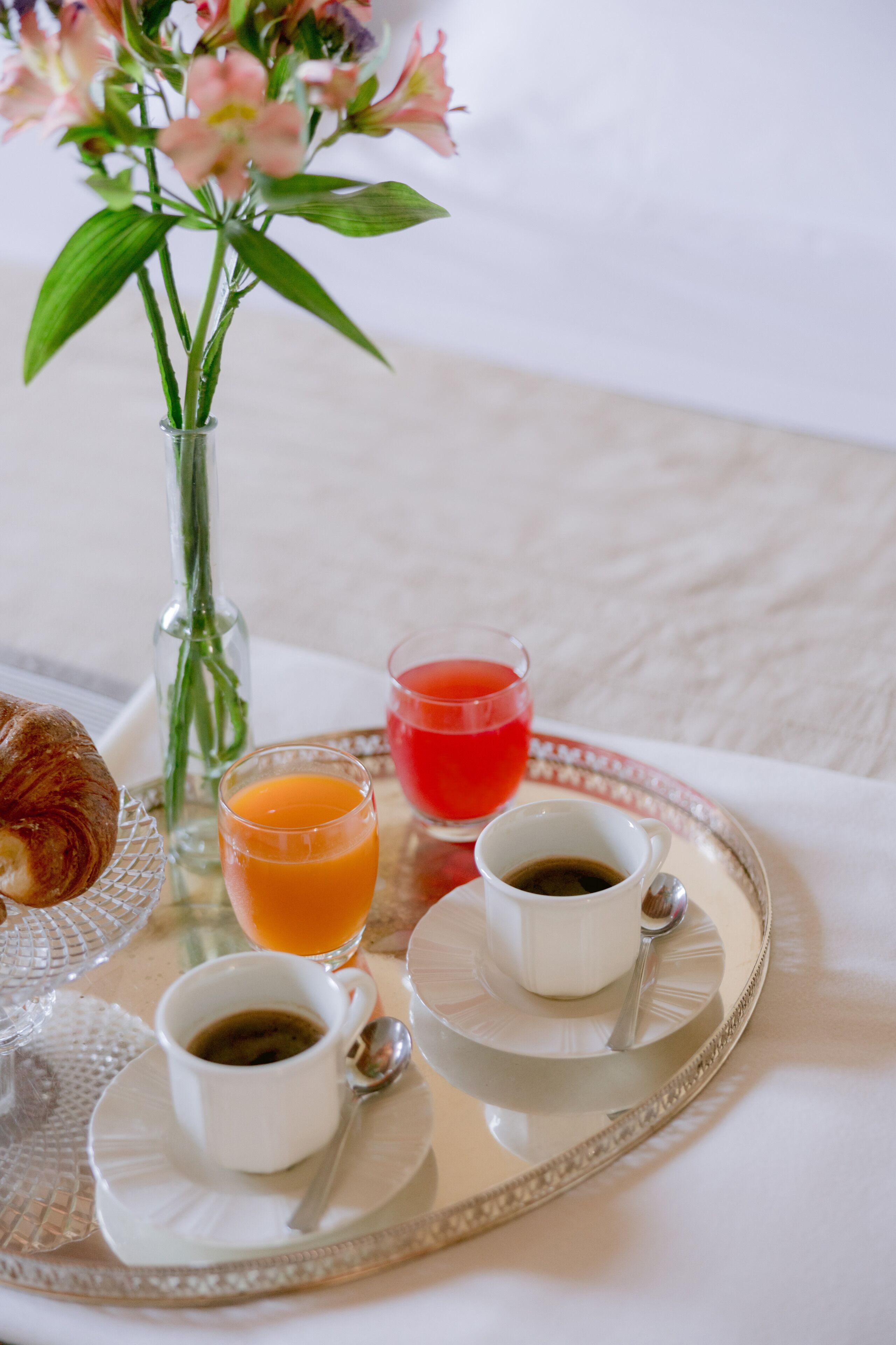 Ein Frühstücksbrett mit Croissant, Orangensaft, rotem Saft und Kaffee auf einem Tisch mit Blumen.