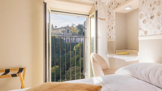A bedroom with a large window, a bed, a bedside table, and a view of a building and trees. - B&B La Rocca (Caprarola)