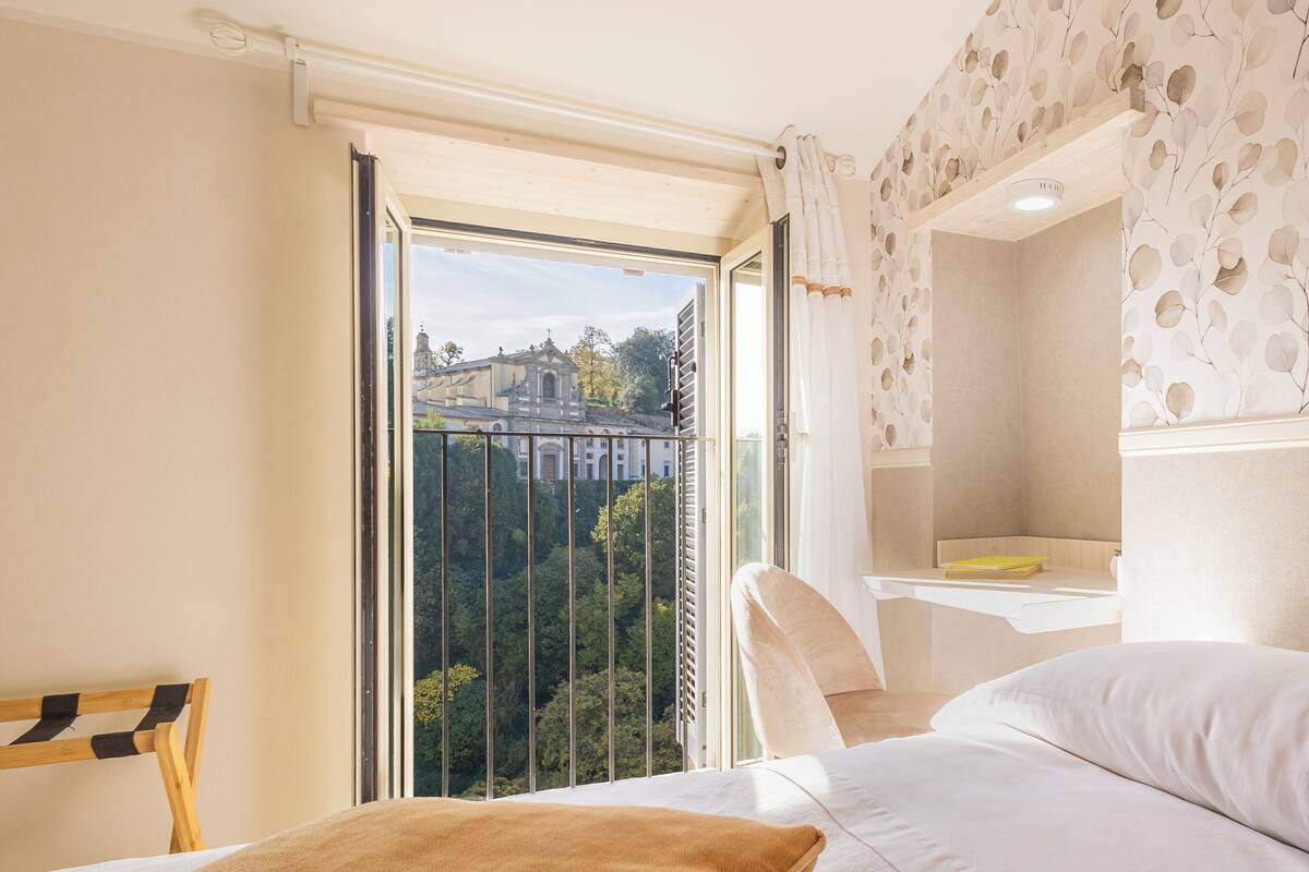 A bedroom with a large window, a bed, a bedside table, and a view of a building and trees.