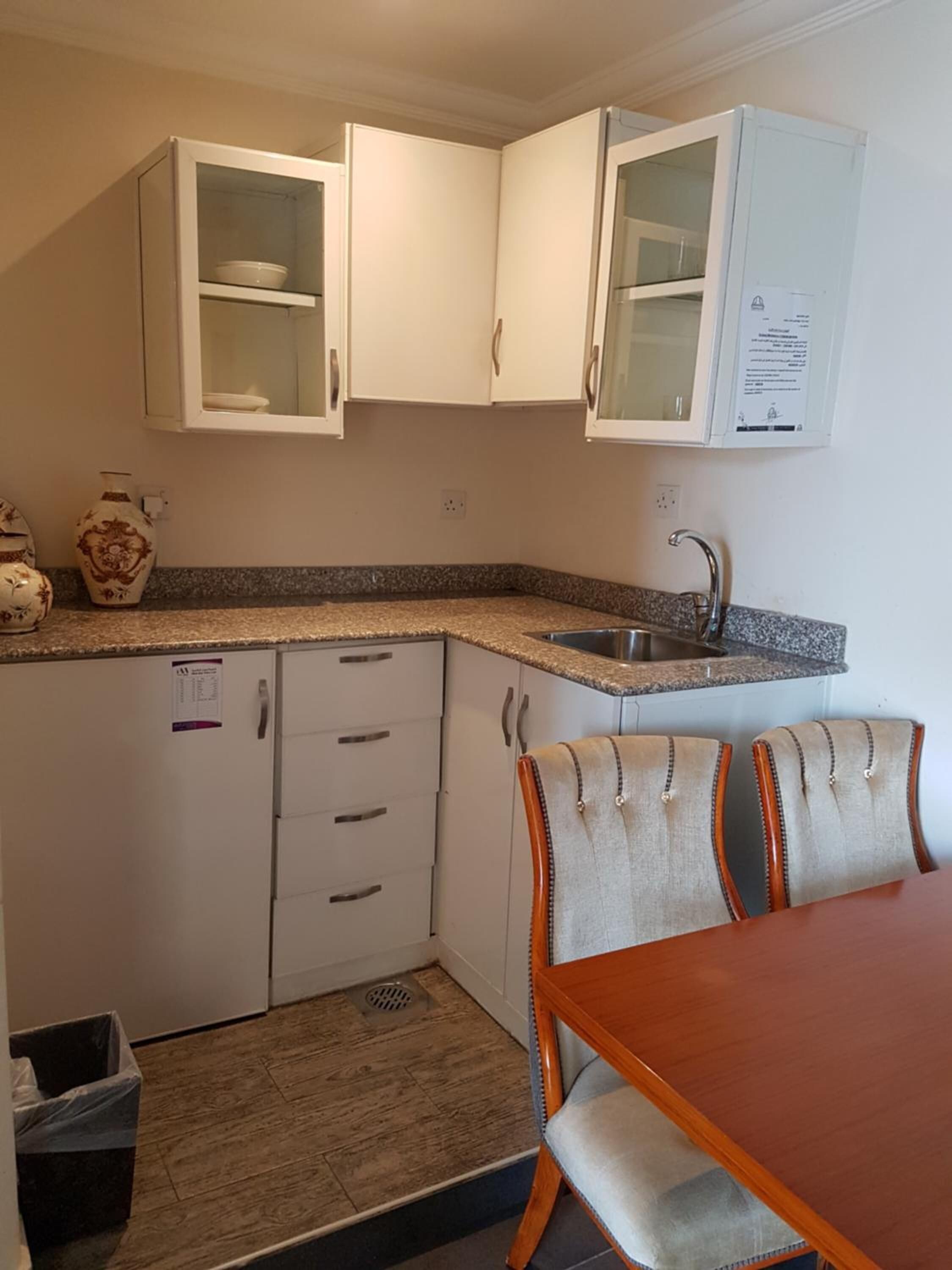 Cocina compacta con armarios blancos, encimera de granito, un lavabo y comedor con mesa de madera y sillas tapizadas.