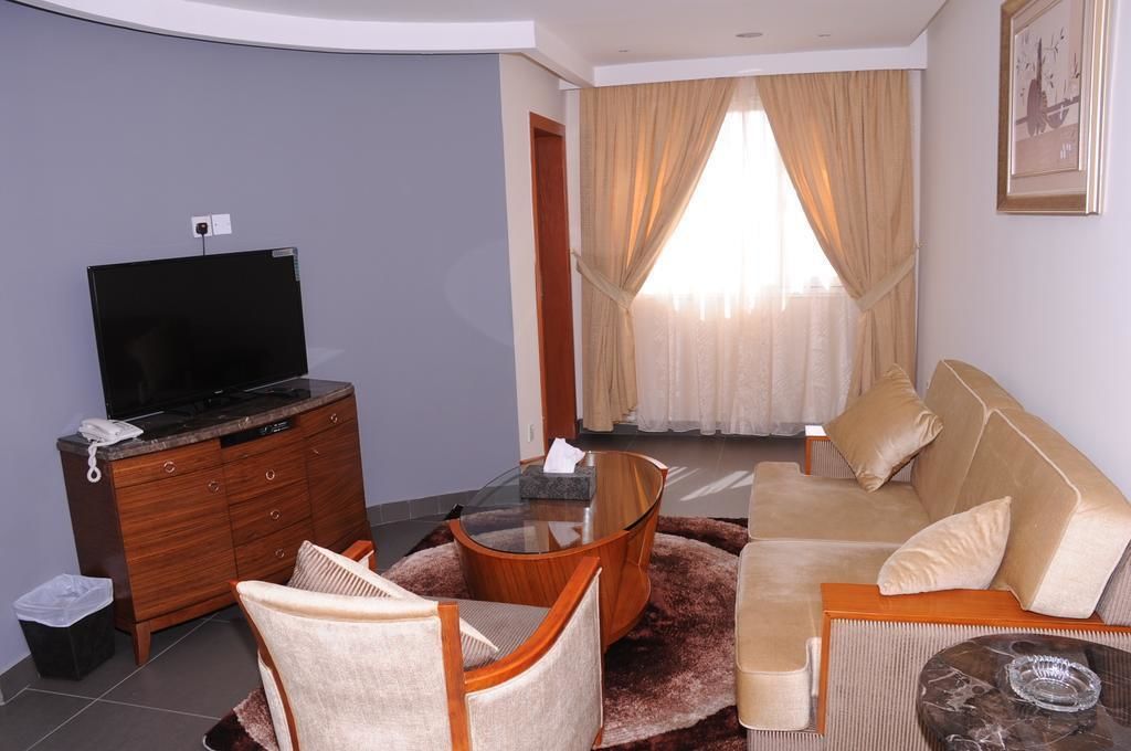 A living room with a flat-screen TV on a wooden cabinet, a beige sofa, a glass coffee table, and a patterned rug.