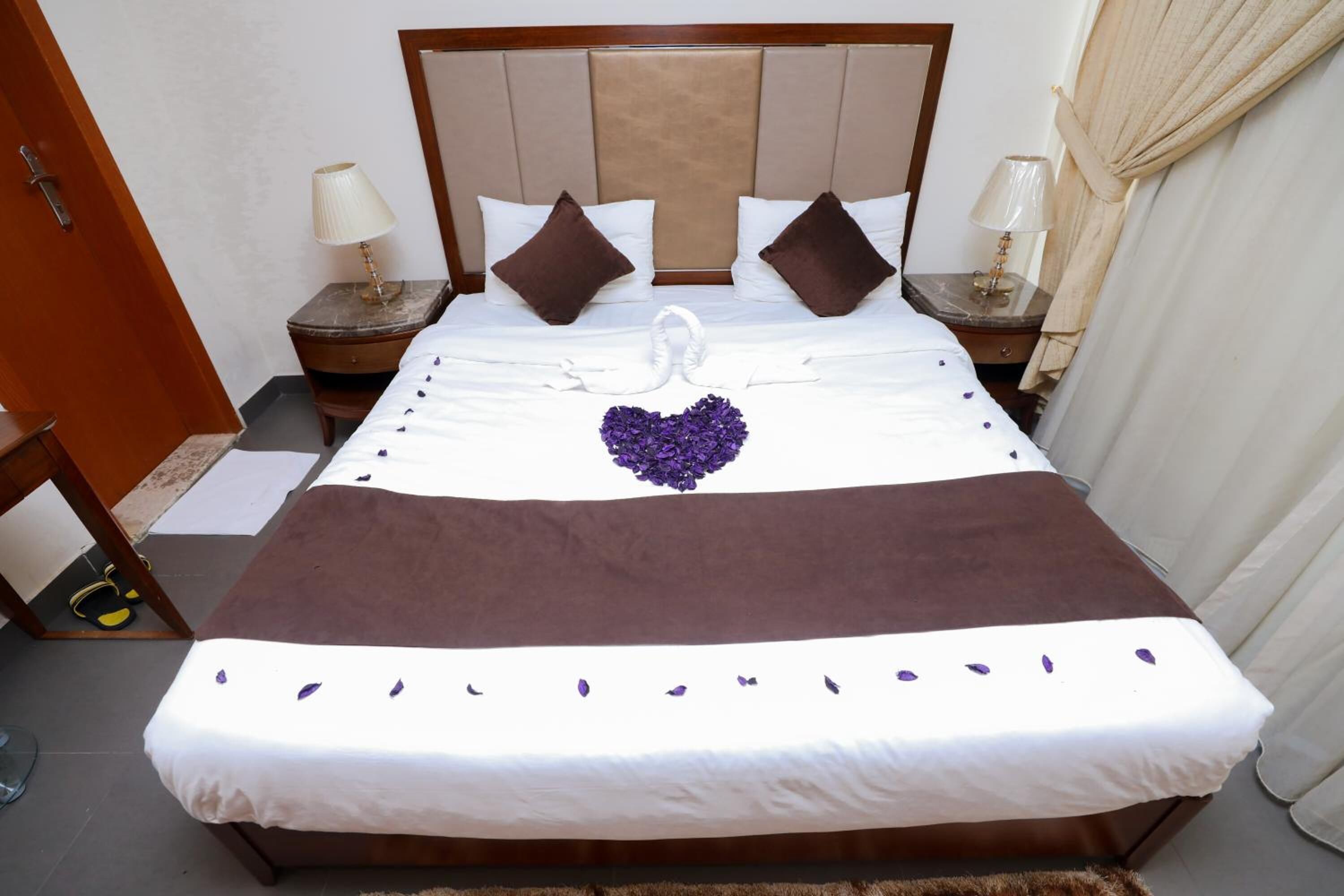 Cama bem arrumada com cabeceira marrom, roupa de cama branca e flores roxas decorativas.
