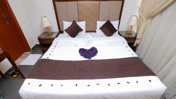 Cama bem arrumada com cabeceira marrom, roupa de cama branca e flores roxas decorativas.
