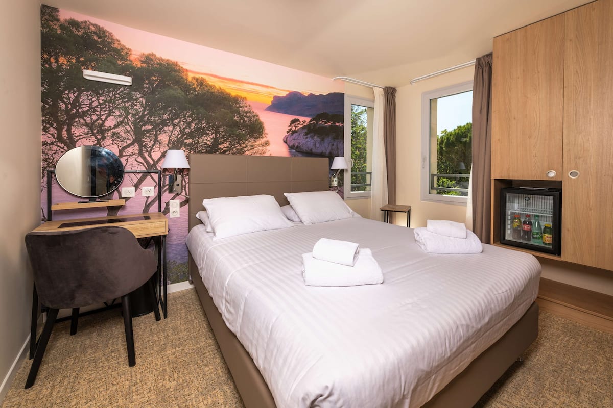 Une chambre d’hôtel avec un grand lit, un bureau avec une chaise, un miroir, un téléviseur fixé au mur et une fenêtre avec des rideaux.