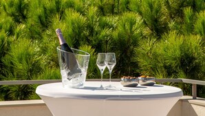 Una mesa con una botella de champán, dos copas y fuentes de comida en un balcón con vistas a un castillo y follaje.