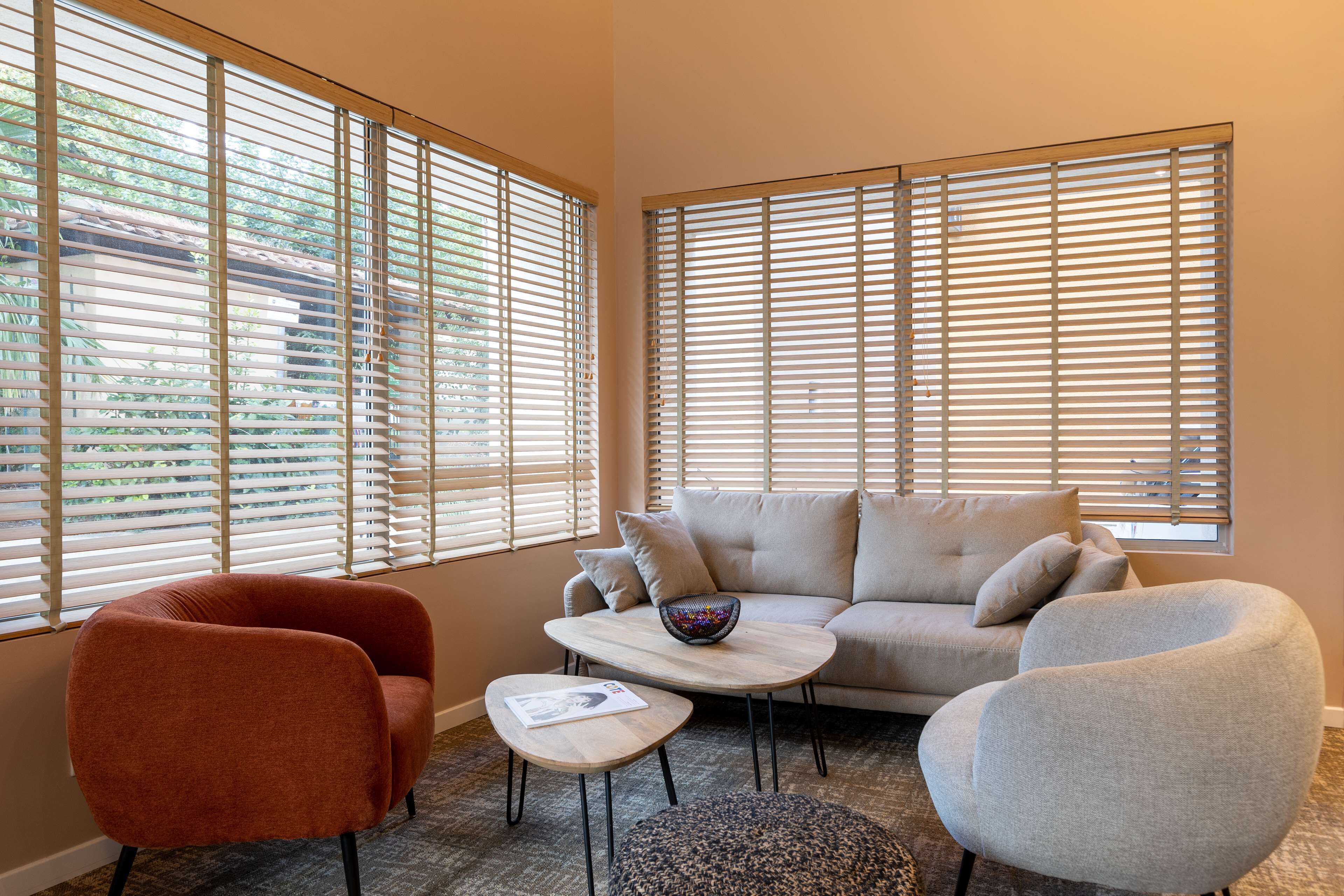 A living room with a sofa, two armchairs, a coffee table, and a small table with a bowl. Large windows with blinds are visible.