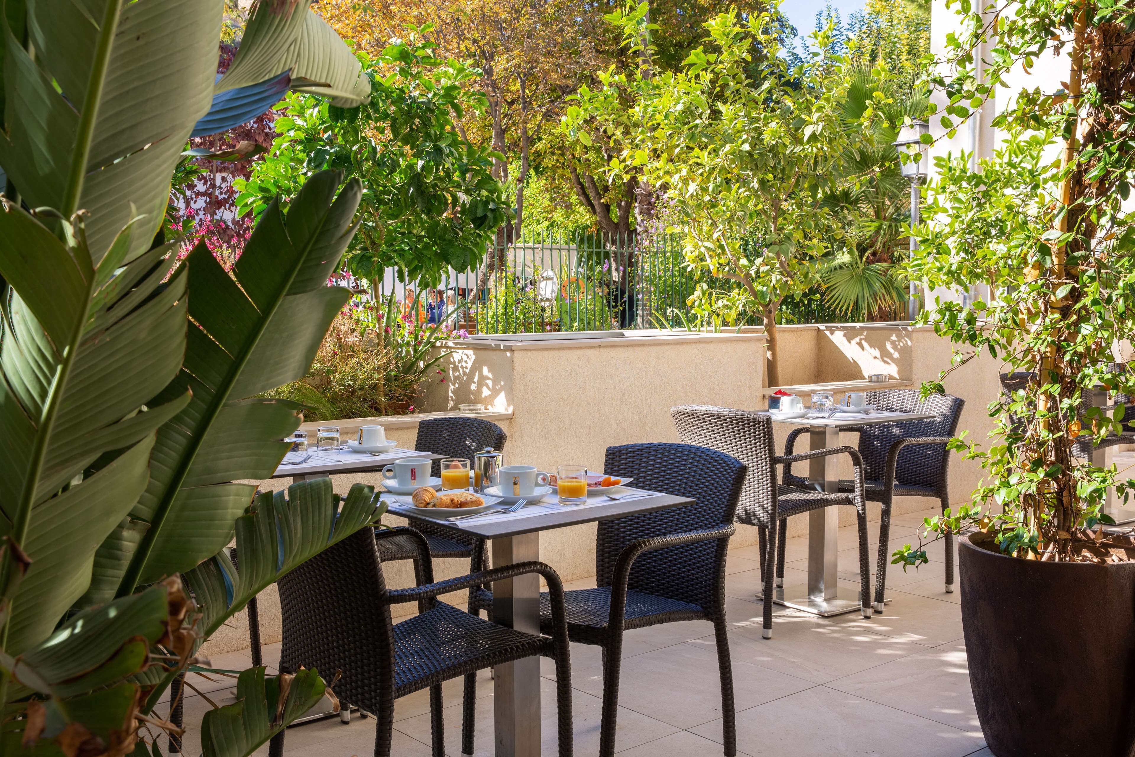 An outdoor café with tables and chairs, surrounded by greenery and a fence.