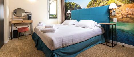 Een hotelkamer met een bed, een bureau met spiegel, een stoel en uitzicht op een kustlandschap.