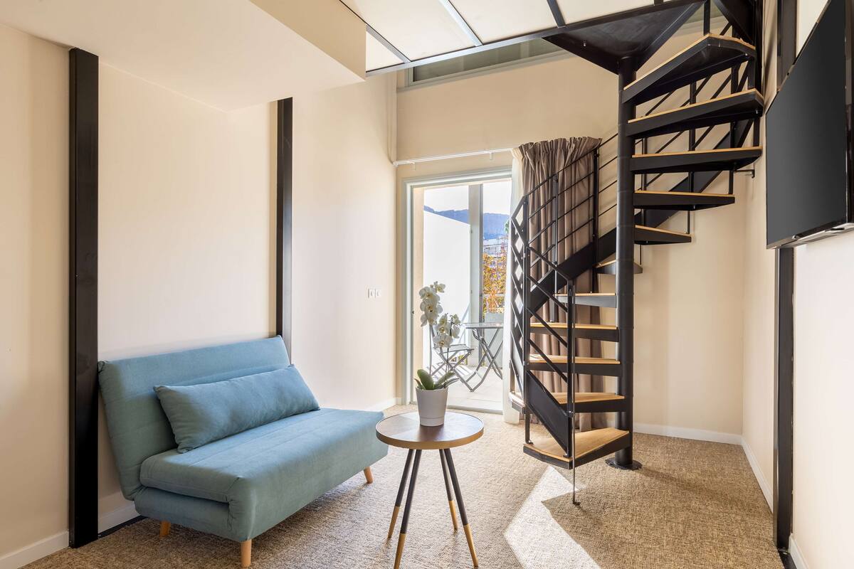 Un salon moderne avec un canapé bleu, une petite table ronde ornée d’une plante et un escalier en colimaçon noir.