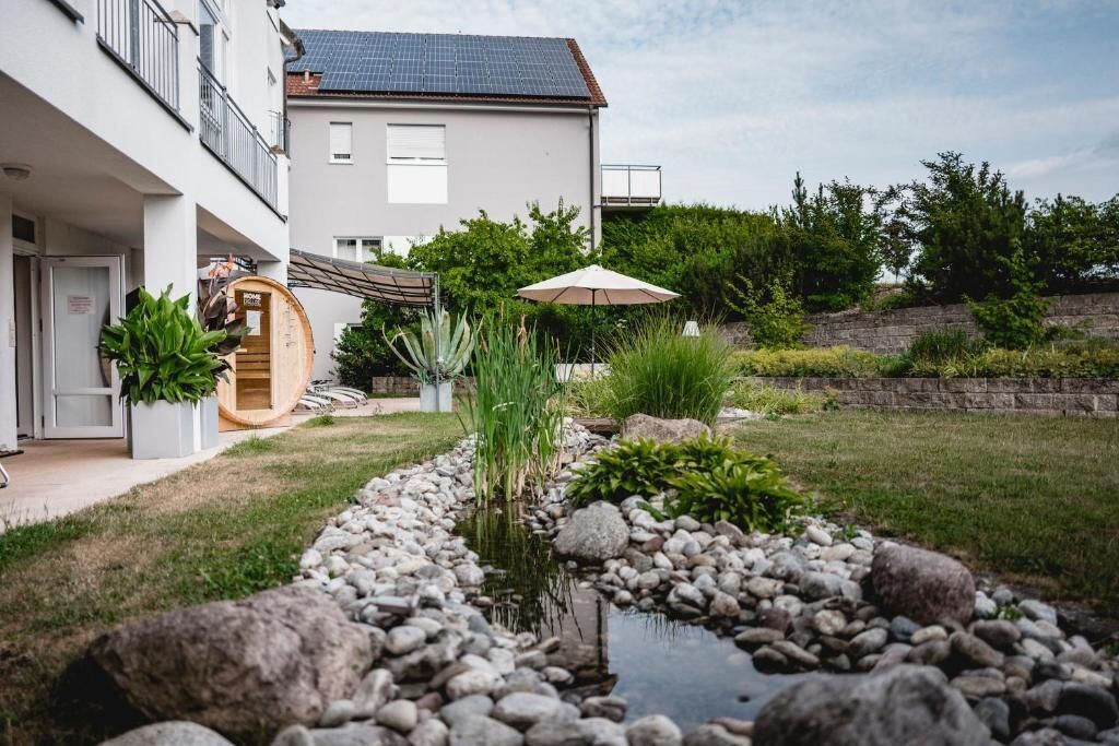 A modern house with a garden featuring a small pond, a wooden barrel, and a patio with an umbrella.