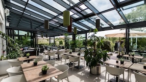 Un café en plein air avec des tables et des chaises, entouré de plantes en pot et doté d'une structure de toit en verre.