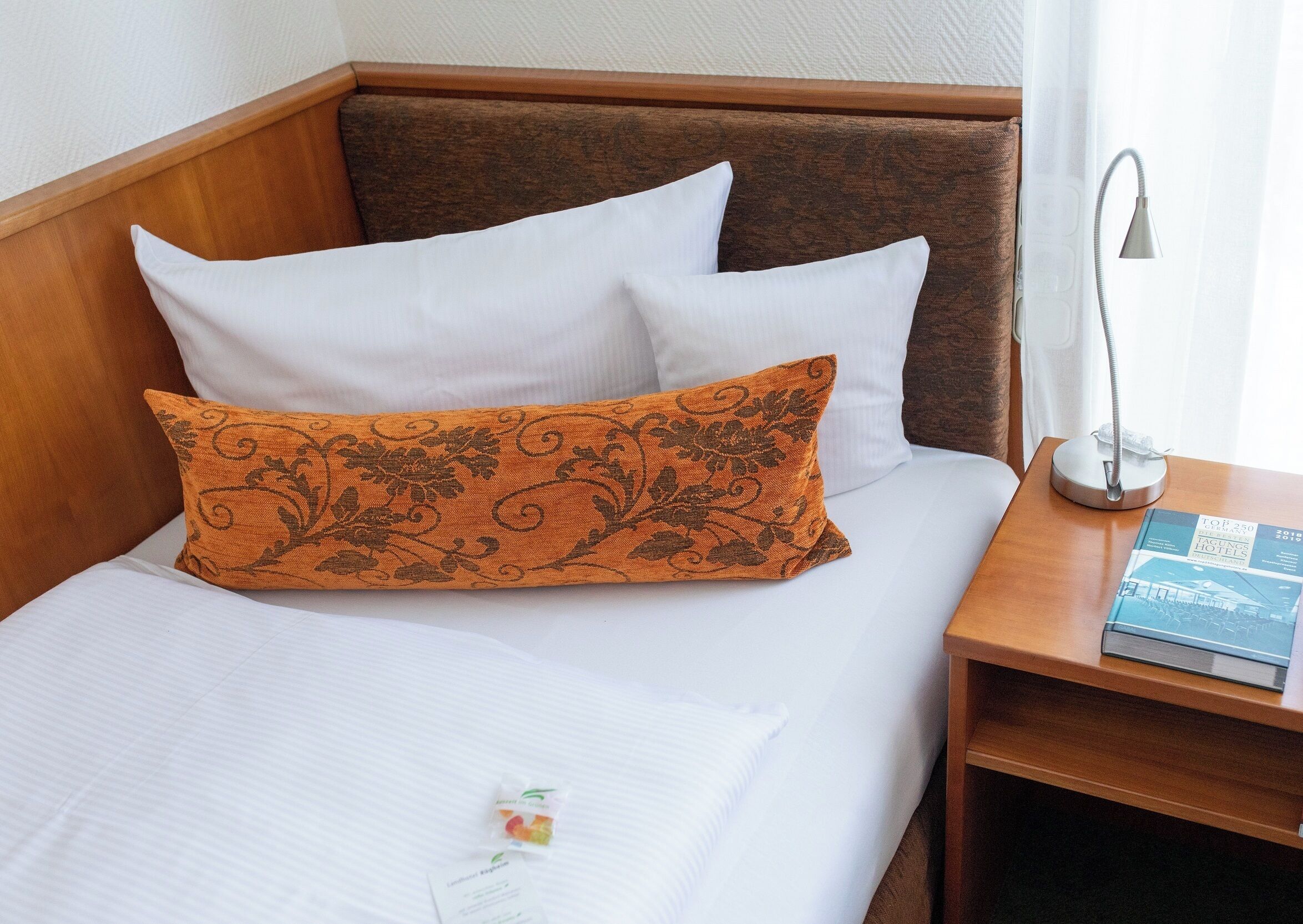 Een netjes opgemaakt bed met witte en oranje kussens, een bedlampje en een boek op een houten nachtkastje.