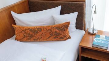 Een netjes opgemaakt bed met witte en oranje kussens, een bedlampje en een boek op een houten nachtkastje.