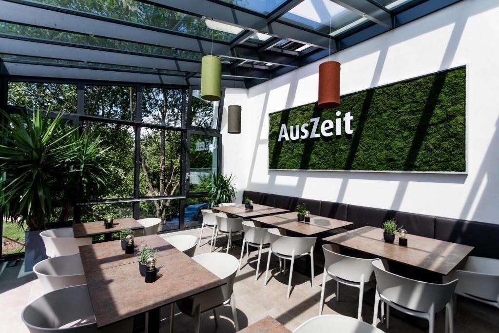 Un área de comedor interior y exterior con mesas y sillas, una gran pared verde con el texto 'AusZeit' y un techo de cristal.