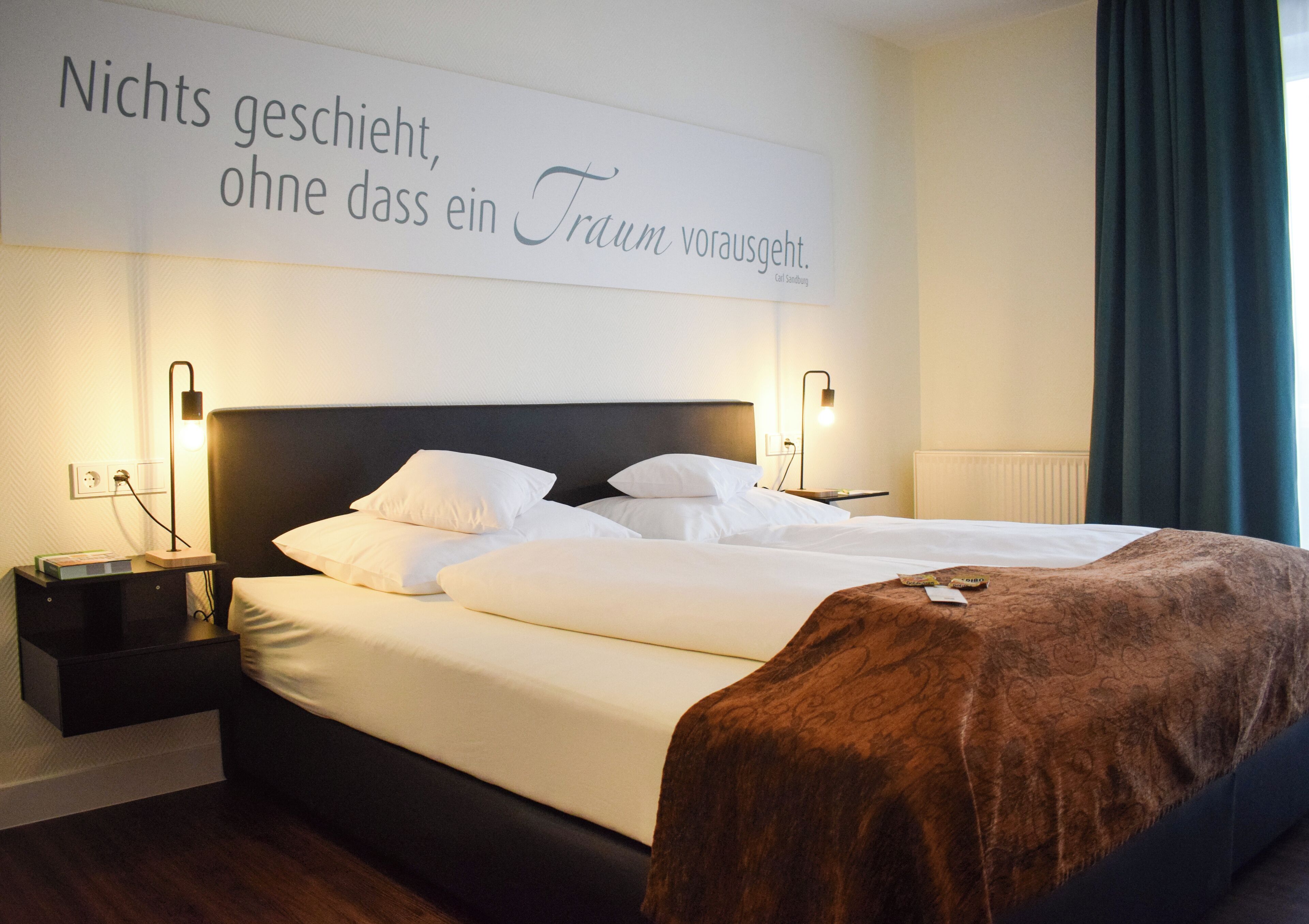 A neatly made bed with white linens and a brown throw blanket, a bedside table with a lamp, and a wall sign with text.