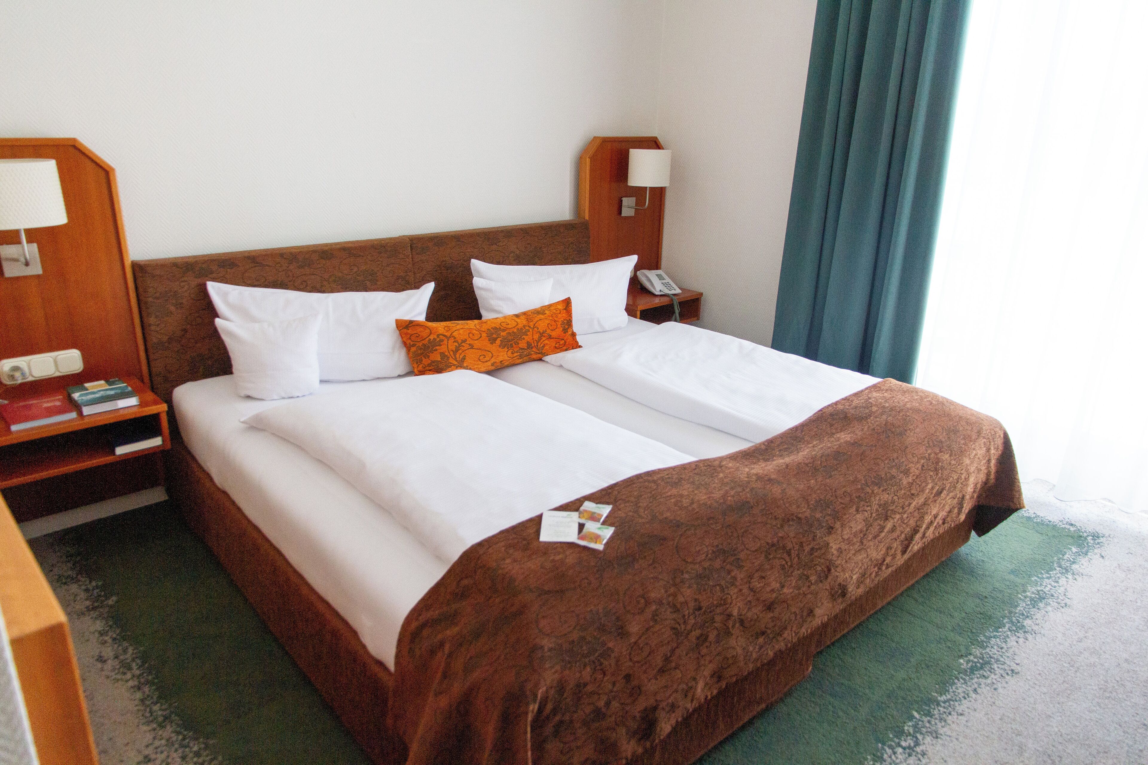 Cama bem arrumada com roupas de cama brancas e castanhas, um cobertor marrom e um travesseiro laranja. Tem uma mesinha de cabeceira com luminária e telefone. O quarto tem cabeceira de madeira e tapete verde.