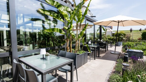 Une terrasse extérieure avec des tables et des chaises en verre, entourée d’une végétation luxuriante et agrémentée d’un grand parasol.