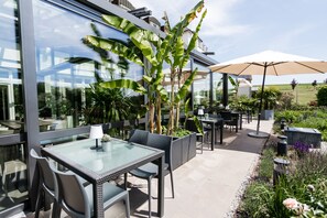 Un patio al aire libre con mesas y sillas de vidrio, rodeado de abundante vegetación y un paraguas grande.