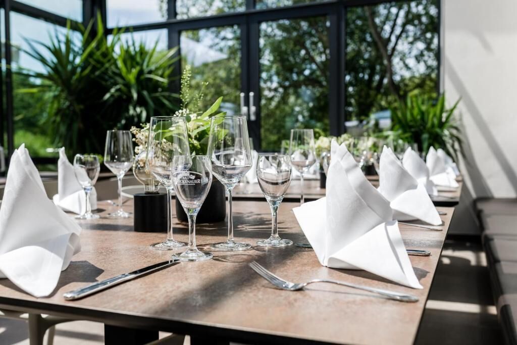 A table set for dining with multiple wine glasses and napkins, set against a backdrop of greenery and a building exterior.