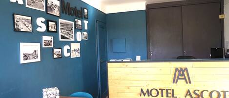 Een receptiegedeelte met een blauwe muur, een houten receptiebalie met de tekst 'Motel Ascot' en enkele zwart-witfoto's aan de wand.